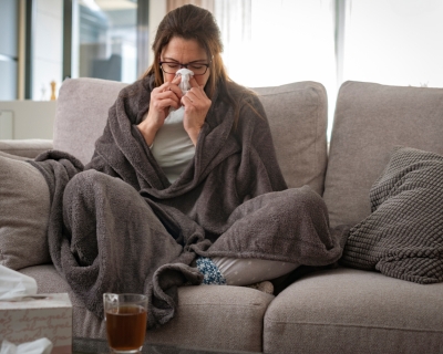 A woman sits on a couch wrapped in a blanket blowing her nose 