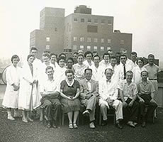 Black and white photo of a group of people, long white labcoats, suits, or dresses gathered in front of a building