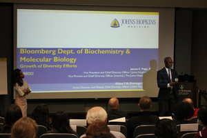 Two people at the front of an auditorium flank a screen with a slider reading Department of Biochemistry and Molecular Biology Growth of Diversity Efforts