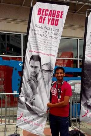 Aravinth stands next to a tall fabric sign reading "Because of you scientists are closer to ending breast cancer" above smaller text and a black and white photo of two people in a lab