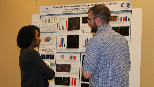 A woman and a man stand talking in front of a research poster with graphs and microscopy images.