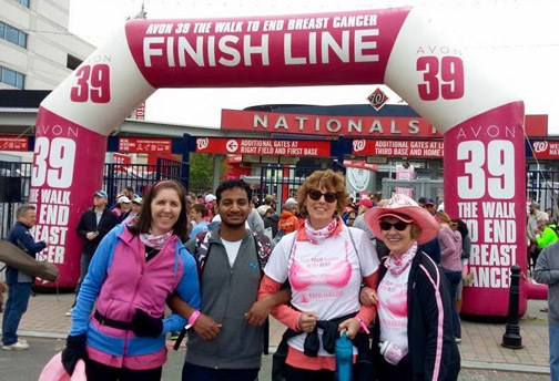 Aravinth and three women stand smiling, arms linked, in front of a large pink and white inflated arch reading "Avon 39 Walk to End Breast Cancer Finish Line"