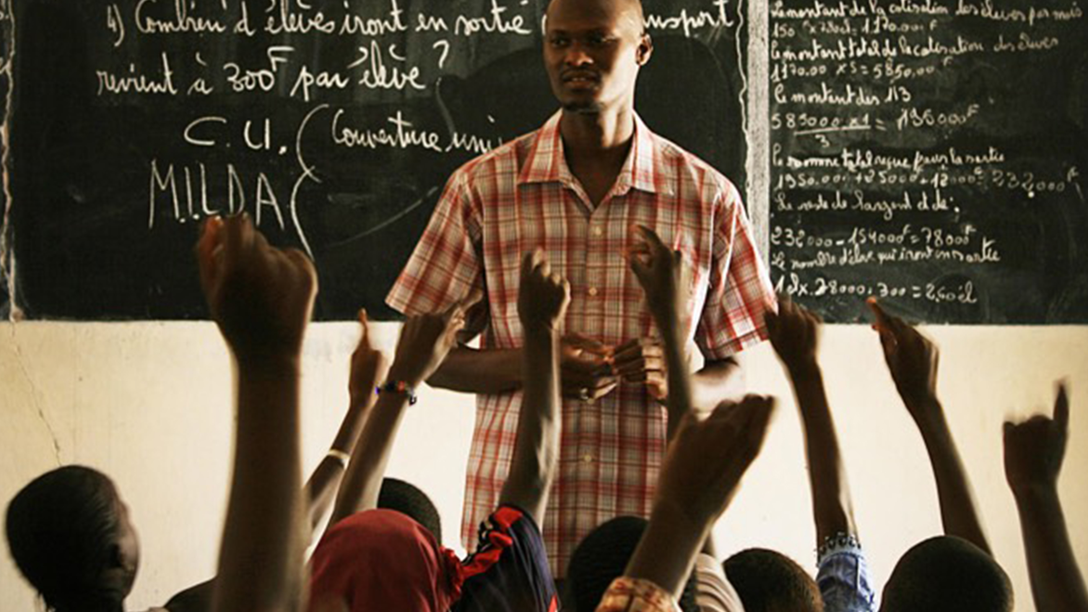 Teacher in front of classroom, with students raising their hands