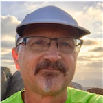 Georg E. Matt, PhD, a man with a light complexion and a goatee, wearing eyeglasses, a blue cap, and a green shirt, photographed outdoors in front of a cloudy sky with sunlight peaking out