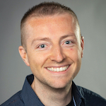 Eric Crosbie, a smiling man with a light complexion, short blondish-brown hair, blue eyes, and wearing a navy blue collared shirt with a black crewneck t-shirt underneath, posed against a grey backdrop