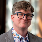 Ryan Kennedy, PhD, a man with a light complexion and wavy light brown hair, wearing black eyeglasses, a colorful collared shirt, and a grey suit jacket, photographed in front of a neutral background