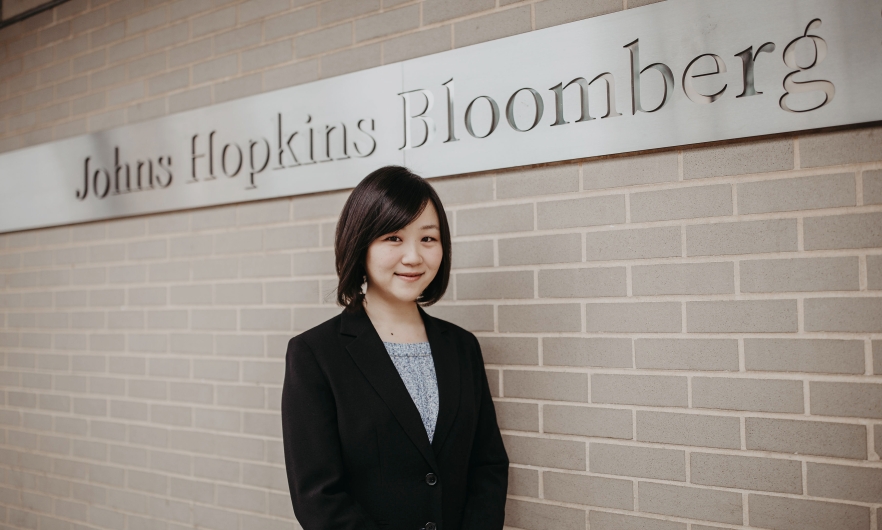 Photo of Megumi "Meg" Ichimiya standing in front of a brick wall, which features a steel signed engraved with the words "Johns Hopkins Bloomberg"