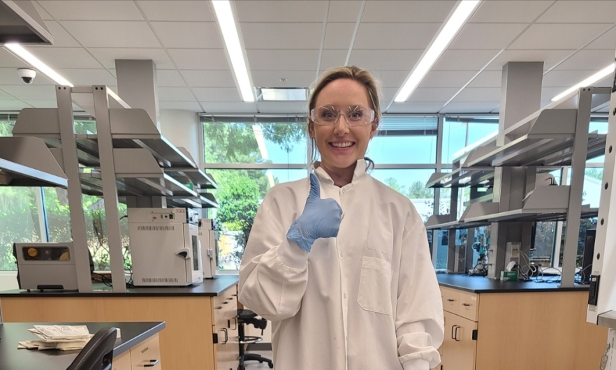 Woman in protective eyewear and a white lab coat gives thumbs up in a lab setting