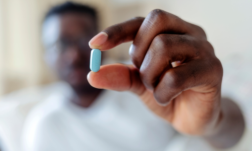 Black man blurred in background holding his hand out which is in focus holding a blue pill