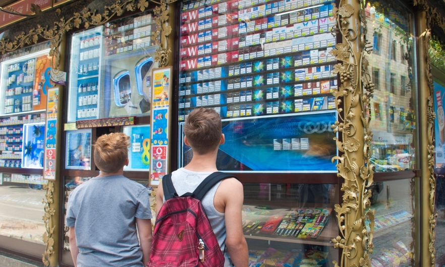 Two young boys stand outside a tobacco vendor in Switzerland, observing a massive display of cigarette products and advertising visible at children's eye level to passersby outside