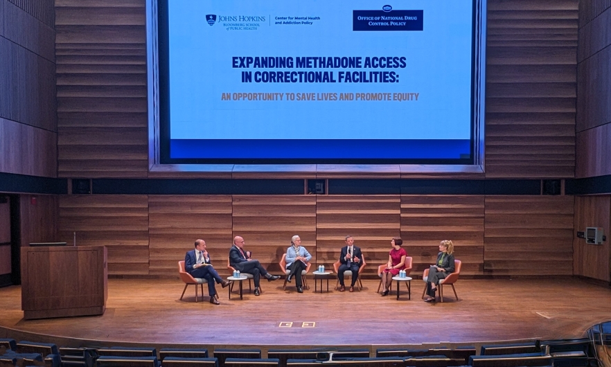 Six adults dressed in business casual attire are seated in a row on stage with a large screen above their heads that says Expanding Methadone Access in Correctional Facilities.
