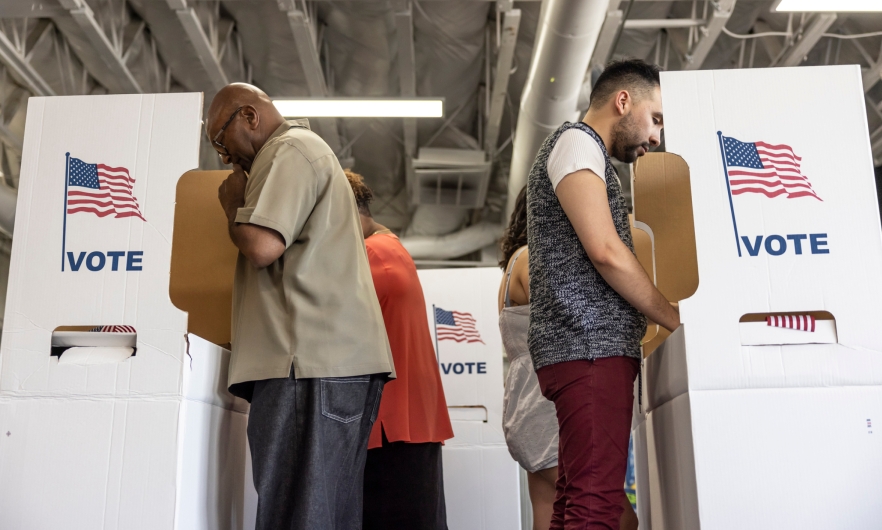 People are voting at cardboard voting stations. The stations have American flags on them and text that says VOTE. 