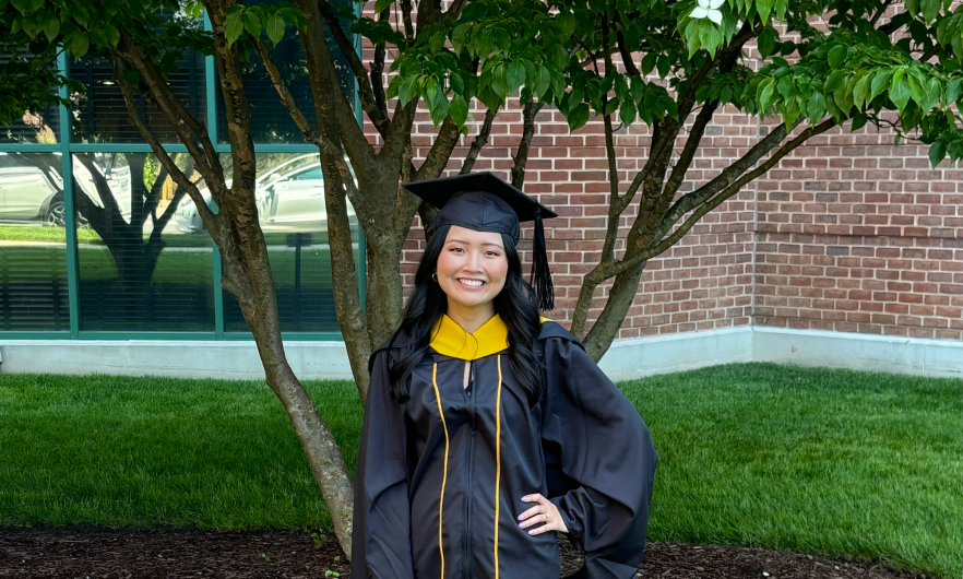 Young woman wearing cap and gown, standing in front of tree.