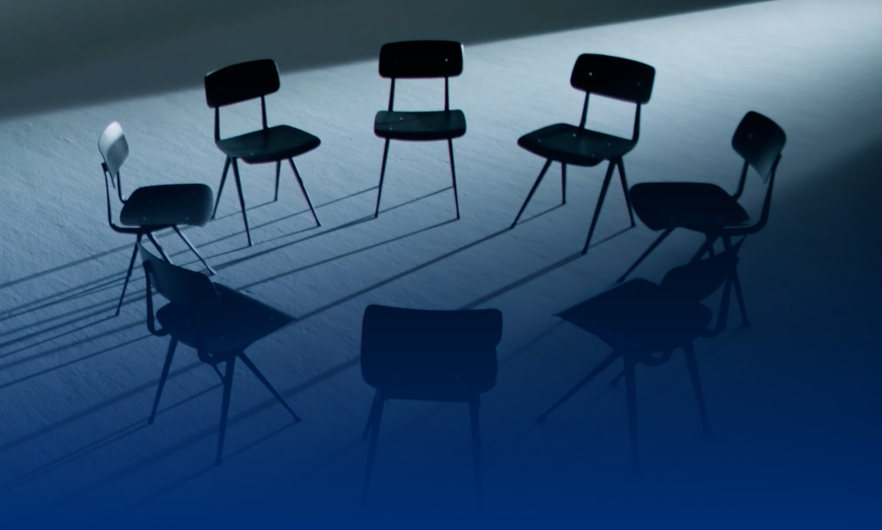 Moody scene of a spotlight casting a shadow across eight folding chairs arranged in a circle in an empty room.