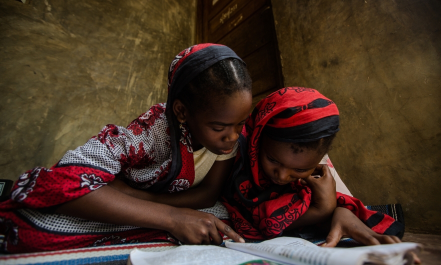 Two girls dressed in red and black garments sit on the floor reading USAID malaria educational materials in Zanzibar, Tanzania. 