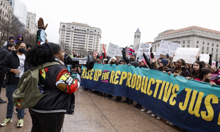 A woman speaks into a microphone, addressing a crowd behind a banner reading &quot;Rise up for Reproductive Justice&quot; at a January 22 protest in Washington, DC. 