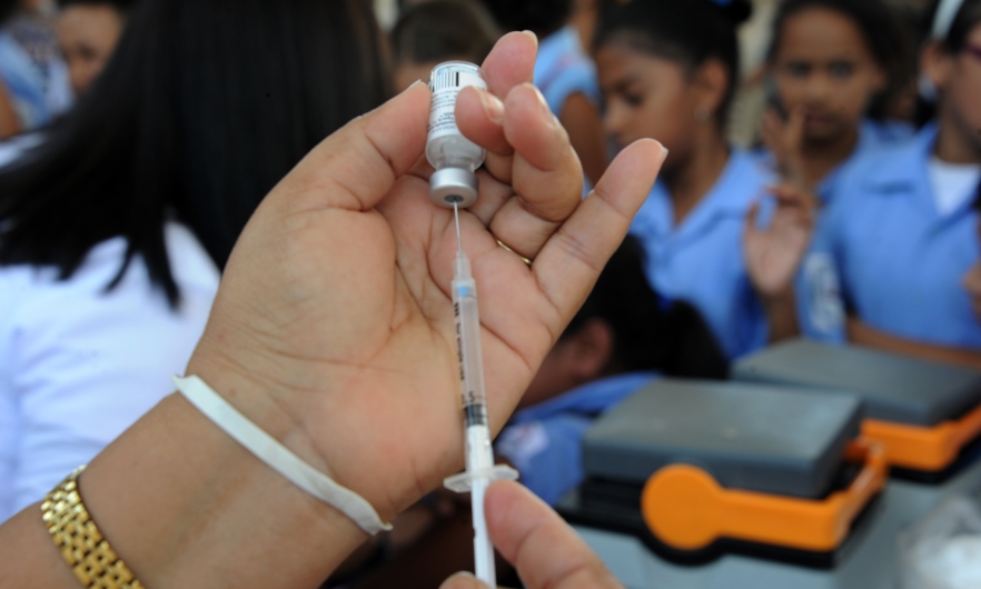 Close-up of a syringe held by a health ministry employee preparing to vaccinate girls against the HPV virus with several girls wearing sky blue uniform shirts in a blurred-out background.