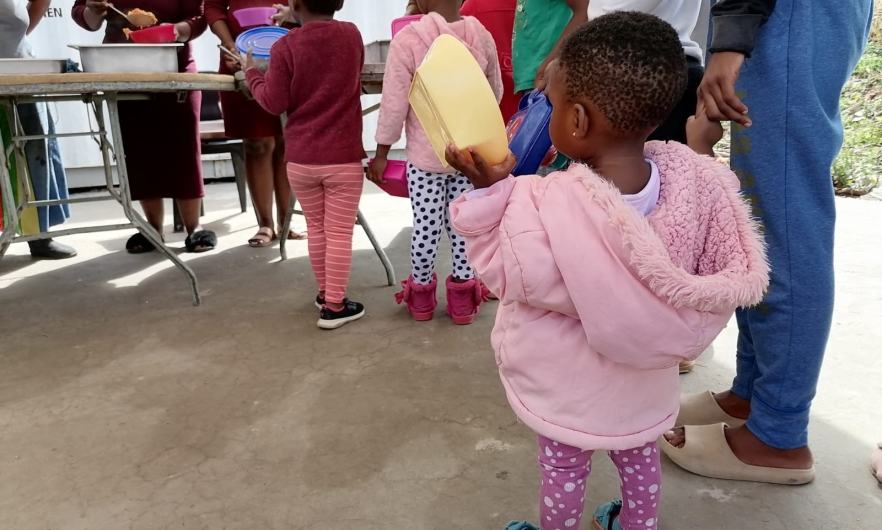 Children, including a toddler in a pink coat holding a yellow lunch container, line up to collect a meal. 