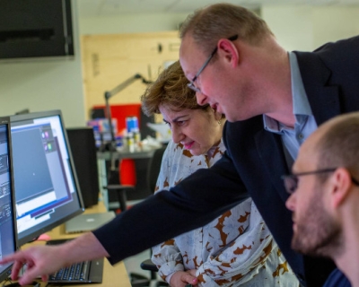 Three people face a desk with a pair of computer monitors on the left. Associate Professor Scott Bailey leans forward, reaching to point at the computer screen. Behind him to the left, Elizabeth Walder stands, looking down at section of the screen that Bailey is pointing at. On the right, in front of Bailey, postdoctoral fellow Evan Worden sits, head turned toward the screen.