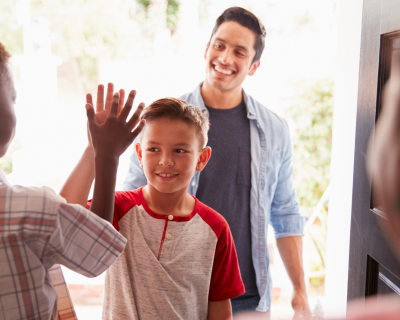 Two children high fiving at the entrance of a house with a father dropping them off