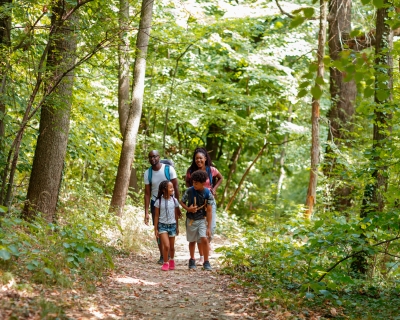 Young African family spends time together, hiking in the woods. They are walking over footpath, carrying backpacks and talking.