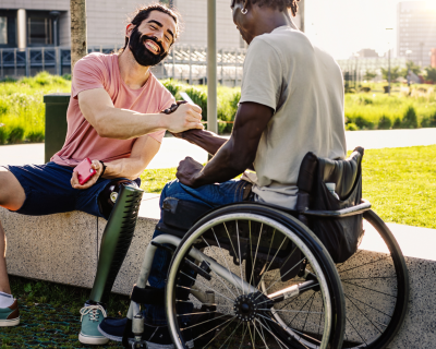 Man in wheelchair shakes hands with a smiling man with a prosthetic leg seated on stone bench outside