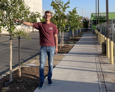 Man in red shirt and jeans standing on a sidewalk with his hand on a tree