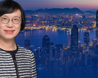 Photo of Dr. Chan, a smiling woman with short parted brown hair, brown eyes, and a light skin complexion, wearing eyeglasses and a black and white striped shirt, posing in front of a photo of the Hong Kong city skyline at night
