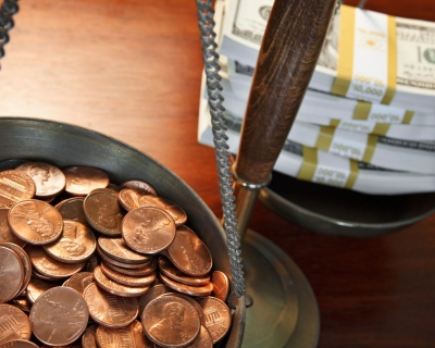 One bowl of an old fashioned weighing scale is full of pennies while the other is full of dollar bill stacks