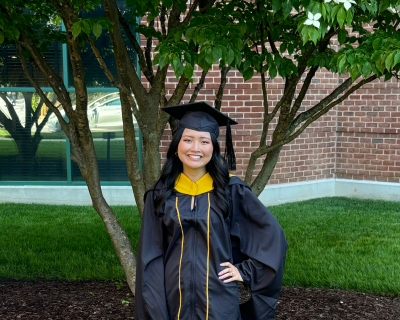 Young woman wearing cap and gown, standing in front of tree.