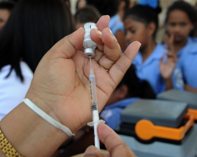 Close-up of a syringe held by a health ministry employee preparing to vaccinate girls against the HPV virus with several girls wearing sky blue uniform shirts in a blurred-out background.