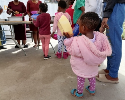 Children, including a toddler in a pink coat holding a yellow lunch container, line up to collect a meal. 