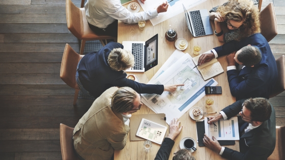 An overhead view of researchers sitting around a table with their laptops. The researchers are engaged in discussion and are actively reviewing documents at the center of the table.