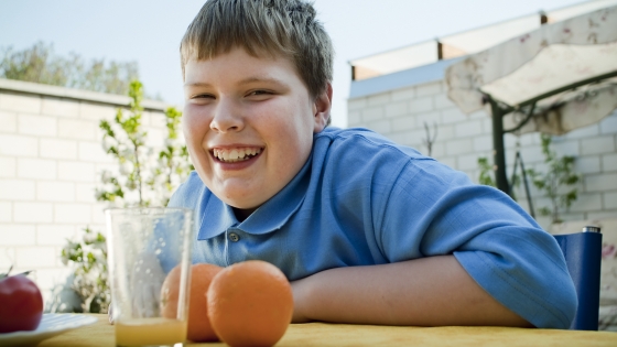 A young teenager (around the age of 13) sits at a table outside in a yard and smiles. At the table next to him are two oranges and an apple, indicating the boy's healthy eating habits.