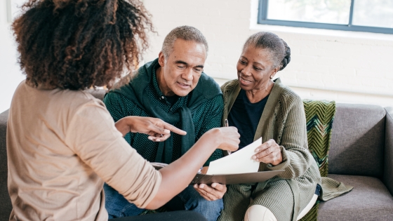 A health professional sits with a middle aged couple and reviews a document summarizing different healthcare options for the couple.