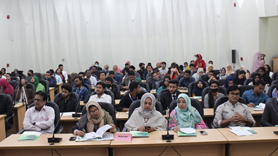 A crowd of people sitting in a large room during a meeting in Bangladesh. Several women with head coverings are in the front row. 