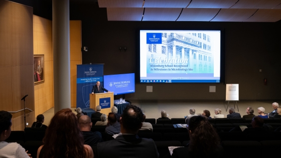 Photo shows a view of Sommer Hall at the Bloomberg School. Arturo Casadevall is to the left behind the podium. Behind him is a Celebration Bloomberg School Recognized As a Milestones in Microbiology Site is on the large screen. Most of the room is filled with people attending the celebration event. 