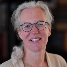 Headshot of Marit van Lenthe, MD, a woman with a light complexion, blue eyes, wavy, grey, shoulder-length hair, wearing a black top, grey jacket, and gold necklace, with a friendly smile on her face