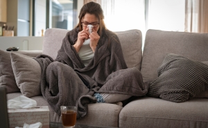 A woman sits on a couch wrapped in a blanket blowing her nose