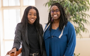 Two black women dressed in business attire smile at the camera as they stand next to each other.