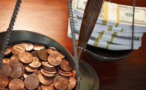 One bowl of an old fashioned weighing scale is full of pennies while the other is full of dollar bill stacks