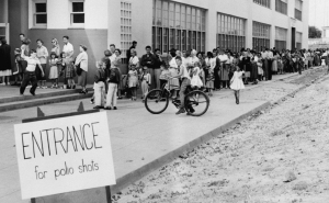 Black and white historical photo from 1955 of families lined up at a school for polio shots. The line wraps around the building.