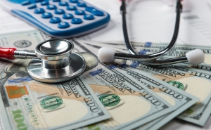 American hundred dollar bills spread out on a desk with a calculator and stethoscope placed on top.