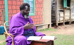 Eliud Wekesa, hands clasped, with a graying beard and wearing a bright purple robe, speaks to visitors outside his home and church compound in Tongaren, Bungoma County, Kenya, on February 29, 2024.
