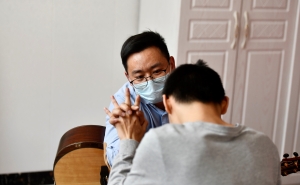 Zhou Pengcheng, wearing a blue surgical mask, clasps hands with his musical therapy patient, a child with short dark hair, shown from the back wearing a long-sleeved grey shirt.