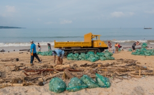 Volunteers remove plastic and other debris from a beach in Kedonganan, Indonesia; dozens of bright green trash bags holding bagged-up debris are piled up around a yellow-orange crawler dump truck.