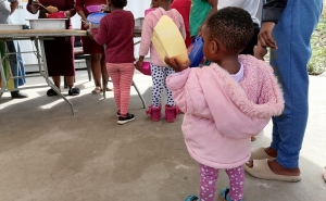 Children, including a toddler in a pink coat holding a yellow lunch container, line up to collect a meal.