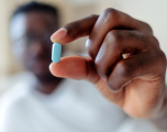 Black man blurred in background holding his hand out which is in focus holding a blue pill